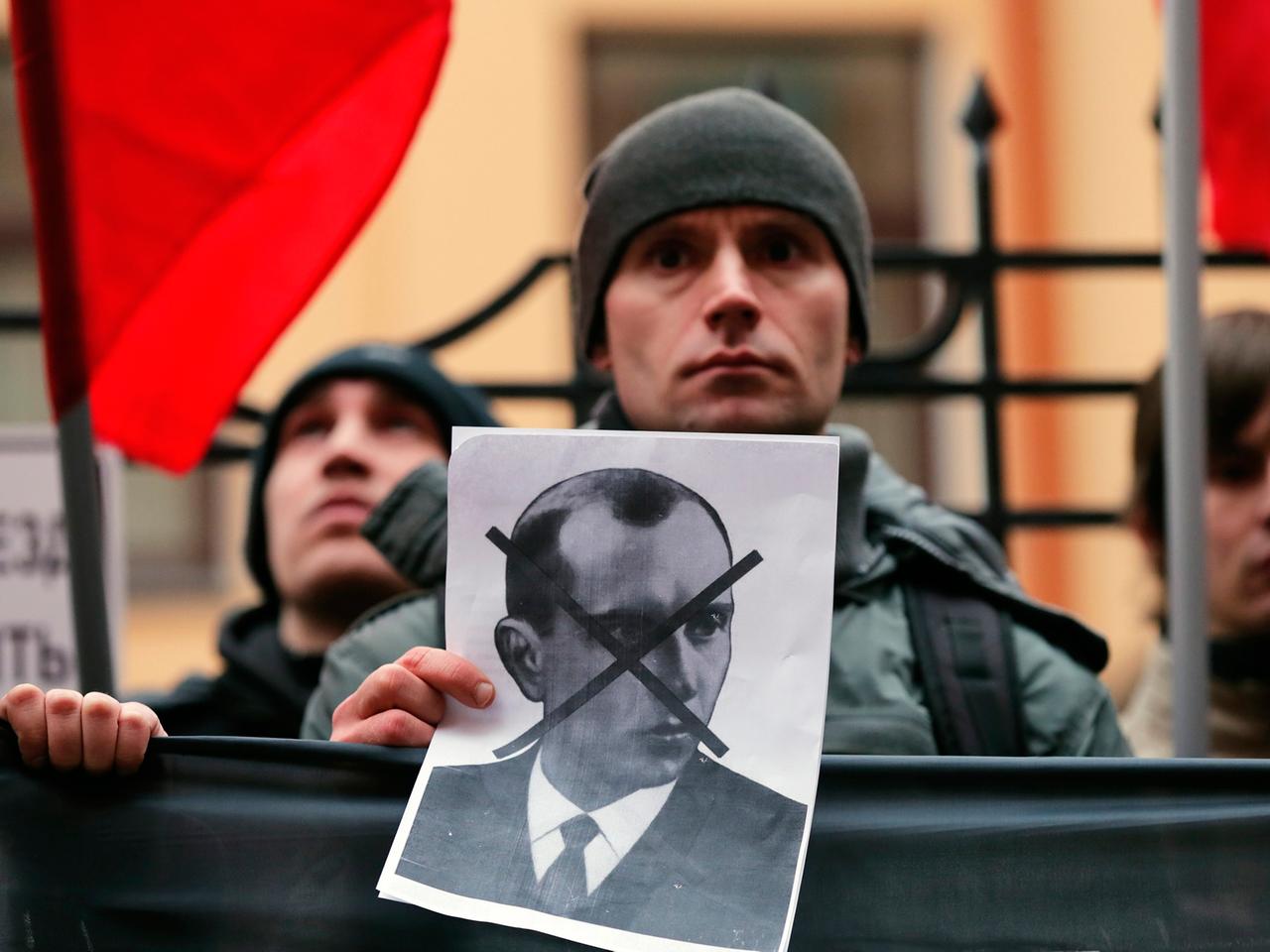 Ein Demonstrant hält ein Schwarz-weiß-Bild von Stepan Bandera in der Hand, dessen Gesicht darauf durchgestrichen ist, im Hintergrund weitere Demonstranten und rote Fahnen