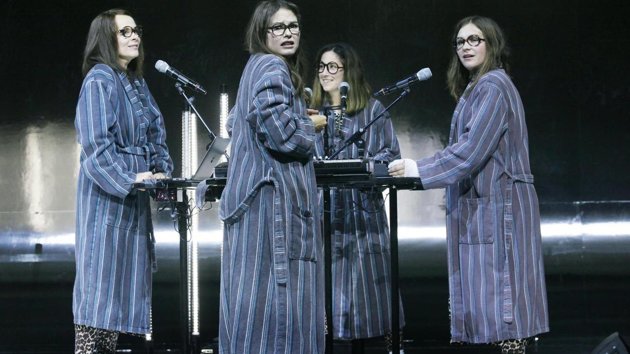 Foto von der Inszenierung: Vier Frauen in Bademänteln stehen im Kreis vor Mikrofonen und Instrumenten. Foto von der Inszenierung: Vier Frauen in Bademänteln stehen im Kreis vor Mikrofonen und Instrumenten.