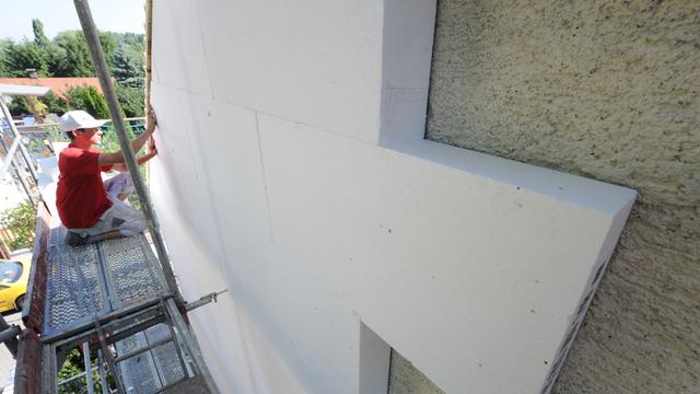 Ein Arbeiter sitzt auf einem Bau-Gerüst und montiert Styropor-Platten an einer Hausfassade.