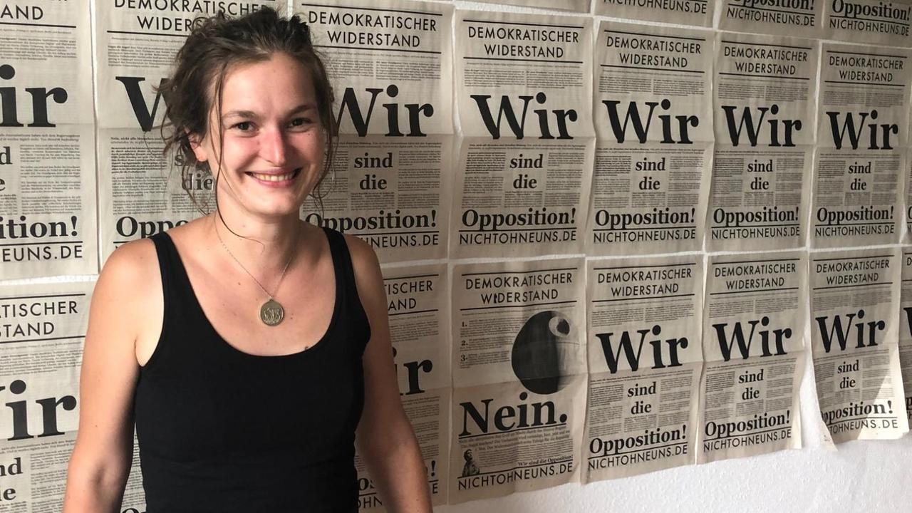 Eine junge Frau steht vor einer Wand mit den Titelseiten der Protestzeitung "Demokratischer Widerstand".
