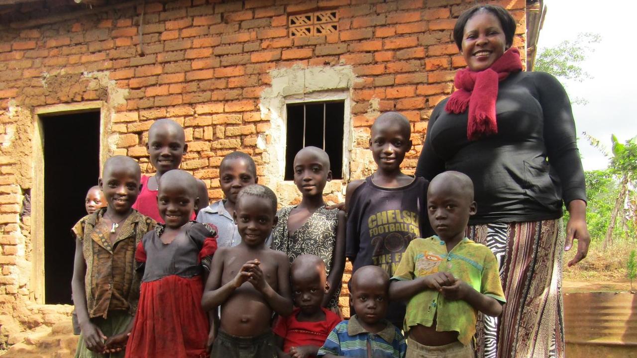 Susan Kigula hilft Kindern von inhaftierten Frauen in Uganda. Sie steht neben einer Gruppe vor einem einfachen Steinhaus. Susan Kigula hilft Kindern von inhaftierten Frauen in Uganda. Sie steht neben einer Gruppe vor einem einfachen Steinhaus.