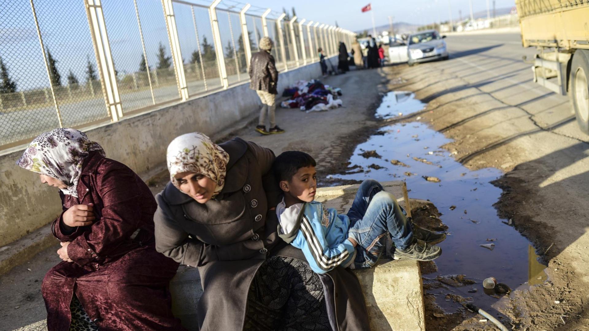 Zwei syrische Frauen und ein Junge warten in der Nähe des Tores von Öncüpınar vor einem Zaun. Zwei syrische Frauen und ein Junge warten in der Nähe des Tores von Öncüpınar vor einem Zaun.