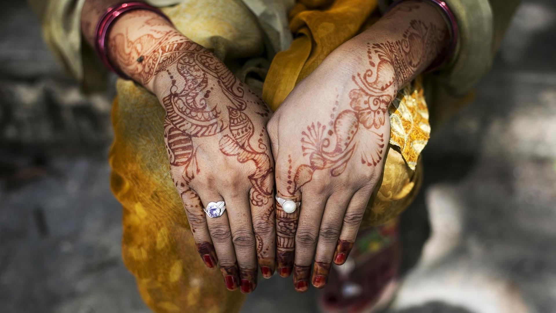 Eine Frau zeigt ihre mit Henna bemalten Handoberflächen. Am linken Zeigefinger trägt sie einen Ring mit einem weißen Stein, am rechten Ringfinger einen Silberring mit lilafarbenem Stein. Eine Frau zeigt ihre mit Henna bemalten Handoberflächen. Am linken Zeigefinger trägt sie einen Ring mit einem weißen Stein, am rechten Ringfinger einen Silberring mit lilafarbenem Stein.