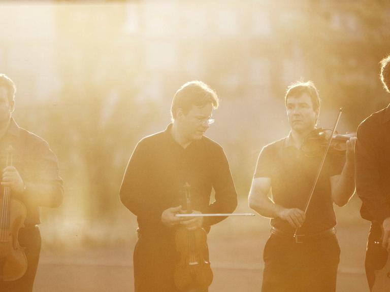Vier Musiker mit Instrumenten stehen in einem Park bei Gegenlicht.