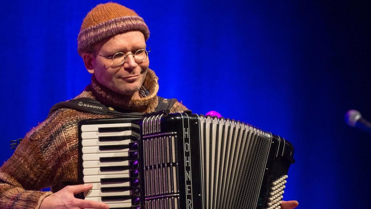 Ein Mann steht mit Wollmütze, kleiner runder Brille, Strickpullover und Akkordeon auf einer Bühne. Ein Mann steht mit Wollmütze, kleiner runder Brille, Strickpullover und Akkordeon auf einer Bühne.