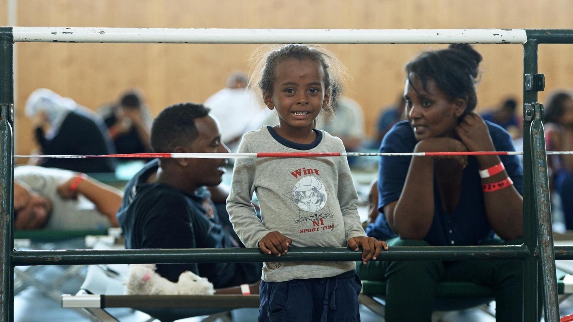 Ein Mädchen aus Eritrea steht neben seiner Mutter vor einem Absperrgitter in einer Turnhalle. Ein Mädchen aus Eritrea steht neben seiner Mutter vor einem Absperrgitter in einer Turnhalle.