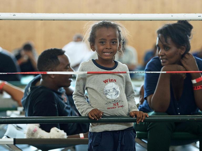 Ein Mädchen aus Eritrea steht neben seiner Mutter vor einem Absperrgitter in einer Turnhalle.