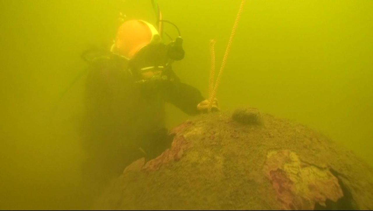 Im trüben Wasser befestigt ein Taucher ein Seil an einer alten, verrosteten Ankertaumine. Im trüben Wasser befestigt ein Taucher ein Seil an einer alten, verrosteten Ankertaumine.