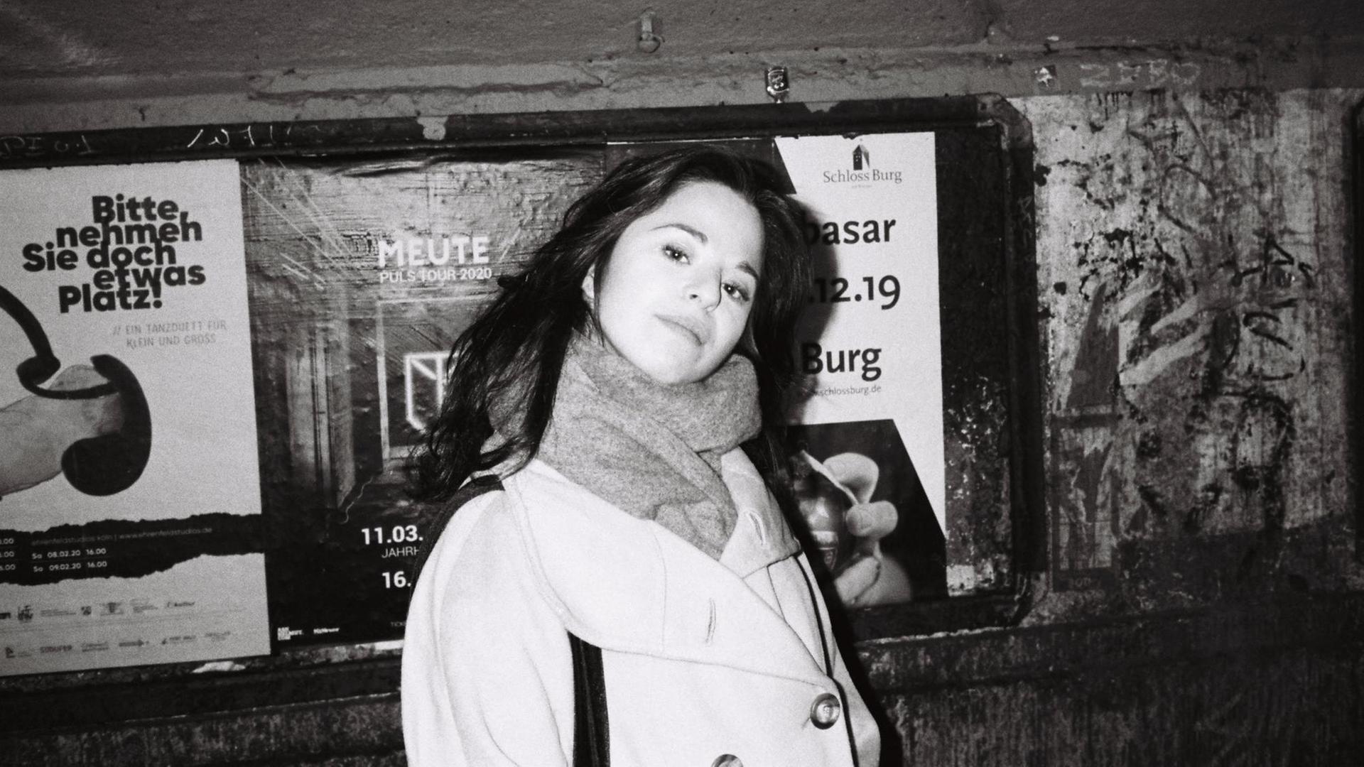 Schwarz-Weiß Aufnahme von Chiara Battaglia, lange dunkle Haare, heller Mantel, vor einer Wand mit Plakaten. Schwarz-Weiß Aufnahme von Chiara Battaglia, lange dunkle Haare, heller Mantel, vor einer Wand mit Plakaten.