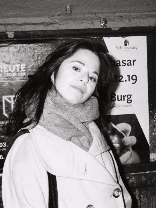 Schwarz-Weiß Aufnahme von Chiara Battaglia, lange dunkle Haare, heller Mantel, vor einer Wand mit Plakaten.