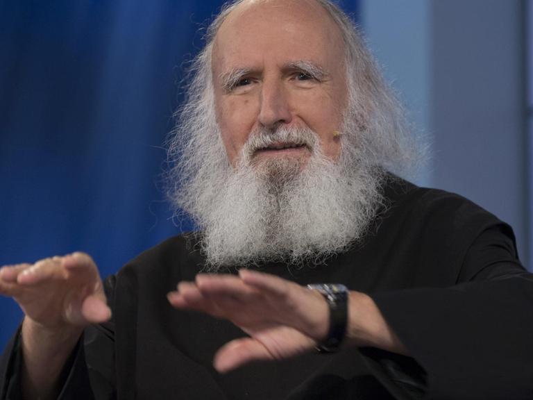 Anselm Grün hat eine Halbglatze, trägt das weiße Haar schulterlang und hat einen langen Vollbart. Er hat die Hände vor der Brust mit den Handflächen nach unten.
