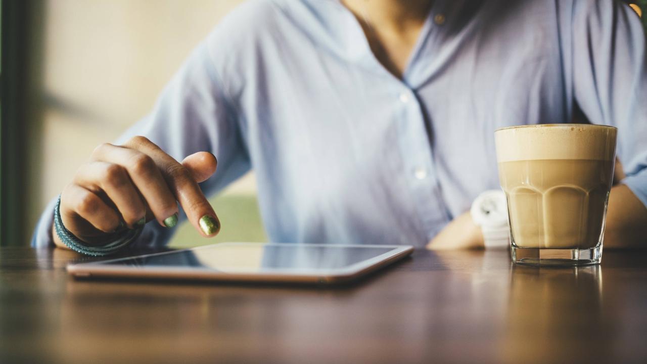 Eine Frau sitzt im Café und nutzt ein Tablet neben dem ein Milchkaffee steht. Eine Frau sitzt im Café und nutzt ein Tablet neben dem ein Milchkaffee steht.