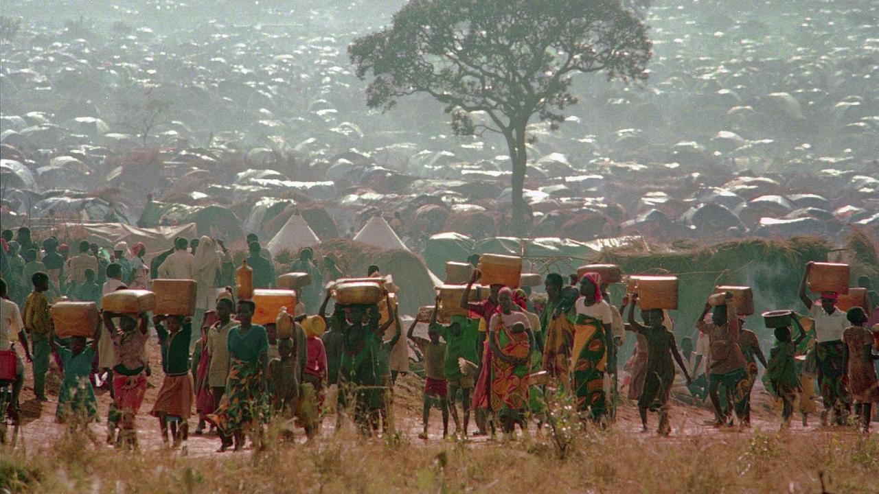 Flüchtlinge, die Wassercontainer tragen vor einem Flüchtlingscamp aus Hütten. Die Flüchtlinge tragen farbenfrohe Kleidung. Zwischen den Hütten steht ein Baum.
