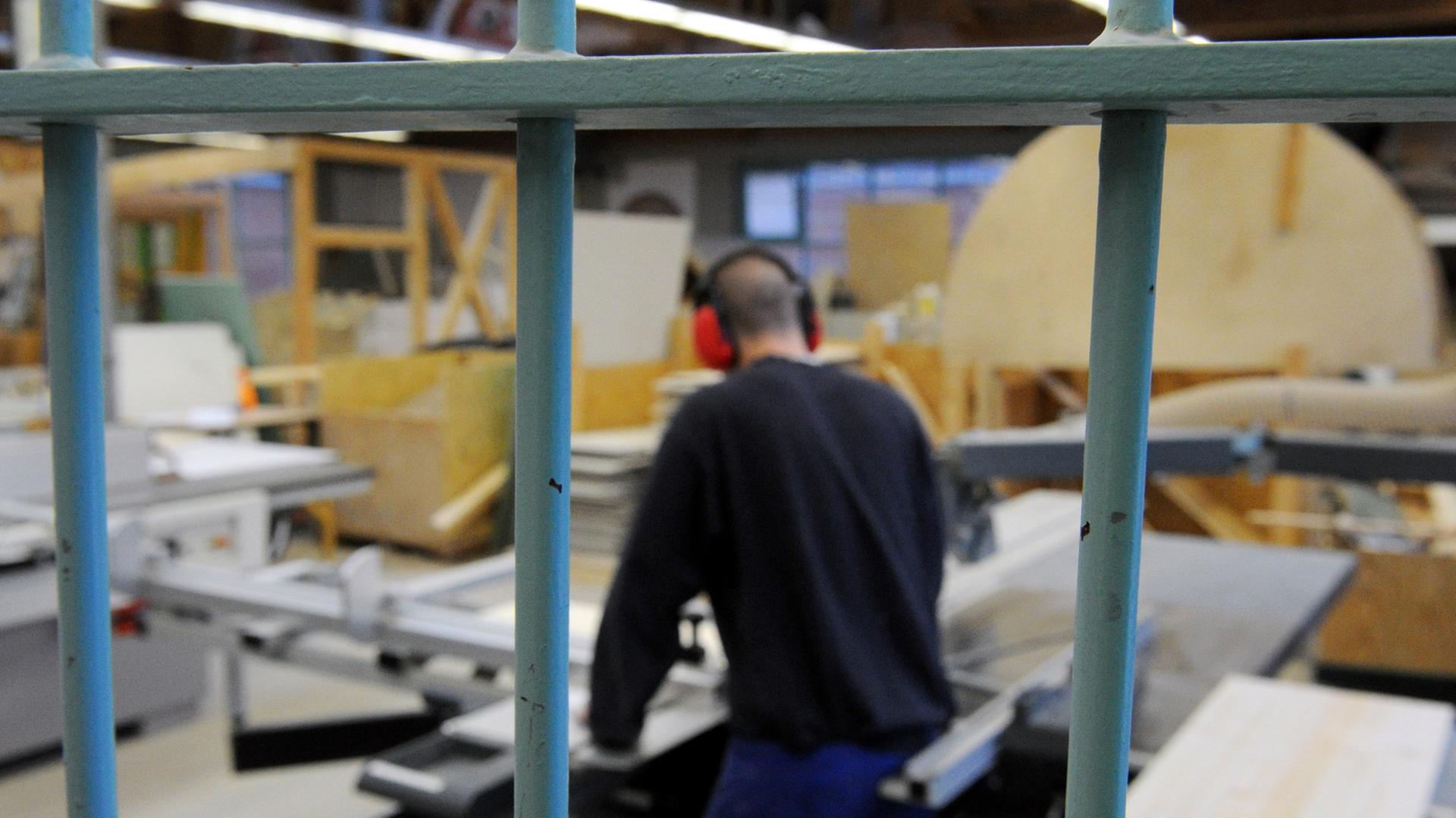 Ein Arbeiter in der Tischlerei einer Justizvollzugsanstalt. Im Vordergrund sind Gitter zu sehen. Ein Arbeiter in der Tischlerei einer Justizvollzugsanstalt. Im Vordergrund sind Gitter zu sehen.