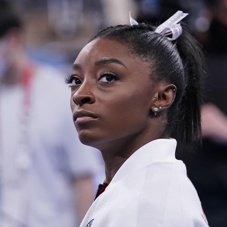 Eine schwarze Athletin im weißem Oberteil blickt ernst an der Kamera vorbei. Es ist die Turnerin Simone Biles.