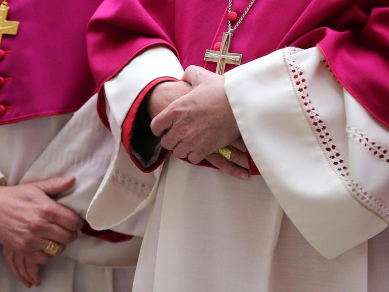 Im Anschnitt fotografierte Würdenträger der katholischen Kirche, sie halten ihre Hände gekreuzt und tragen purpurrote Roben und große Kreuzanhänger um den Hals