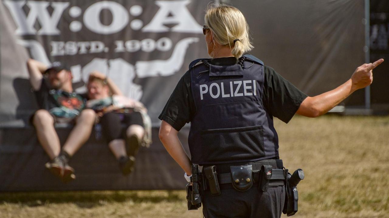Eine Polizistin zeigt mit ihrem Finger beim Wacken-Open-Air in die rechte Richtung. Sie ist von hinten zu sehen. Vor ihr sitzen zwei Menschen auf einem Podest.