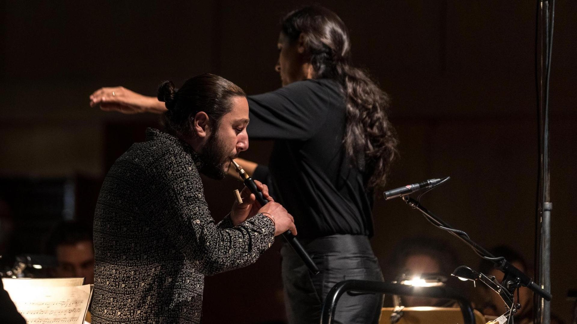 Im Vordergrund ist ein Flötist zu sehen, der inbrünstig in sein Instrument bläst. Im Hintergrund dirigiert eine Frau mit langem Haar Zopf ein unscheinbares Orchester. Im Vordergrund ist ein Flötist zu sehen, der inbrünstig in sein Instrument bläst. Im Hintergrund dirigiert eine Frau mit langem Haar Zopf ein unscheinbares Orchester.