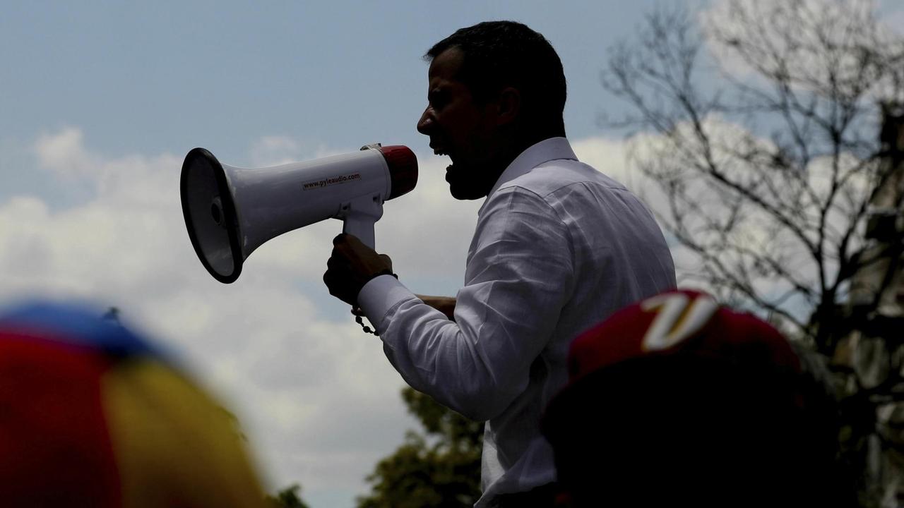 Man sieht Guaidó seitlich im Gegenlicht als schwarze Silhouette. Er hält ein Megafon und ruft etwas hinein. Man sieht Guaidó seitlich im Gegenlicht als schwarze Silhouette. Er hält ein Megafon und ruft etwas hinein.