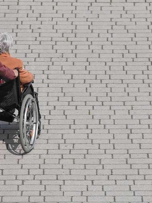 Eine Frau schiebt eine ältere Frau in einem Rollstuhl über einen Gehweg