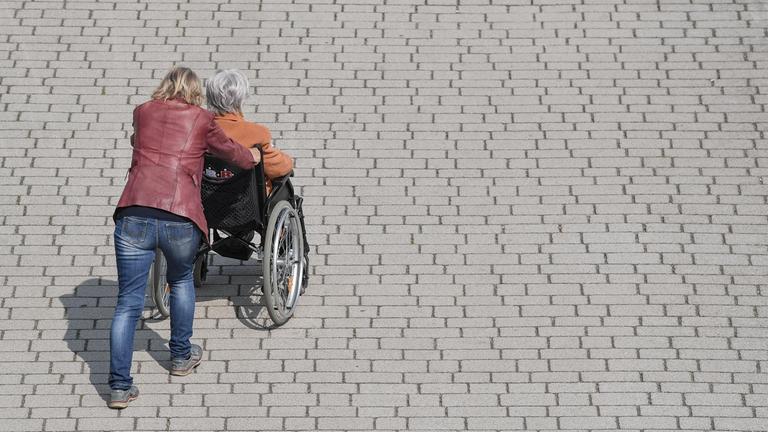 Eine Frau schiebt eine ältere Frau in einem Rollstuhl über einen Gehweg