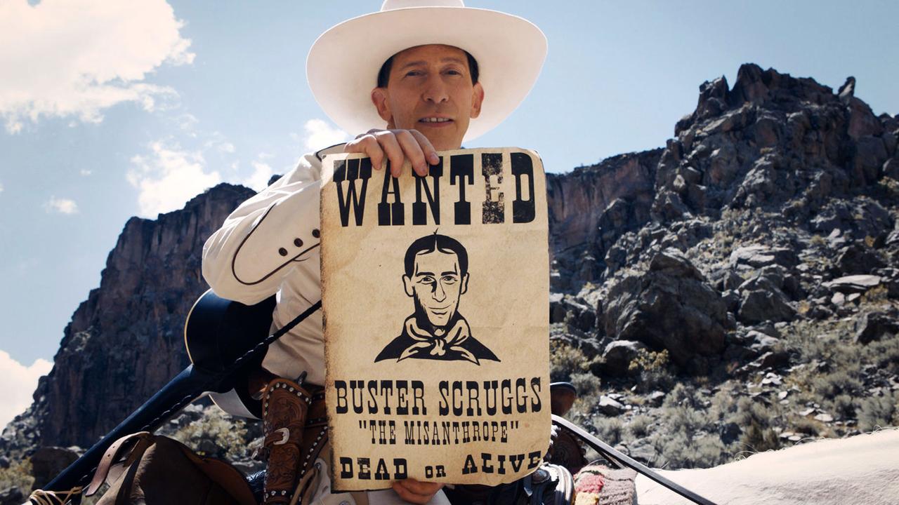 Der Schauspieler Tim Black Nelson steht als Buster Scruggs in einem Wester-Szenario und hält eine Fahndungsrolle mit seinem Gesicht und Name hoch. Der Schauspieler Tim Black Nelson steht als Buster Scruggs in einem Wester-Szenario und hält eine Fahndungsrolle mit seinem Gesicht und Name hoch.