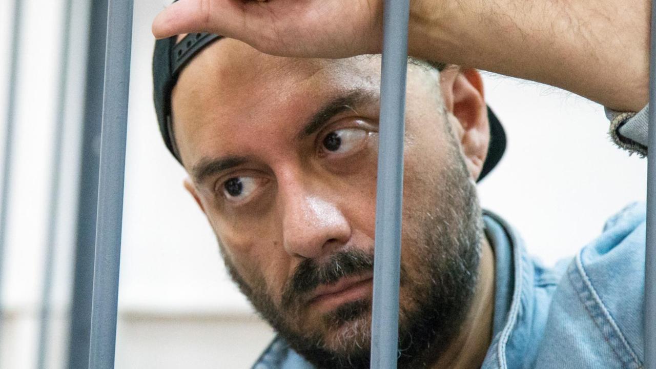 Nahaufnahme von Serebrennikows ernstem Gesicht hinter Gittern. Er lehnt mit einer Hand an den Gitterstäben. Er trägt eine Baseballmütze verkehrt herum und eine blaue Jeansjacke. Nahaufnahme von Serebrennikows ernstem Gesicht hinter Gittern. Er lehnt mit einer Hand an den Gitterstäben. Er trägt eine Baseballmütze verkehrt herum und eine blaue Jeansjacke.