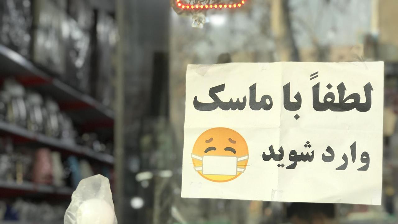 Ein Schild mit Schrift in Farsi und einem gelben Emoji mit weißer Maske. Ein Schild mit Schrift in Farsi und einem gelben Emoji mit weißer Maske.
