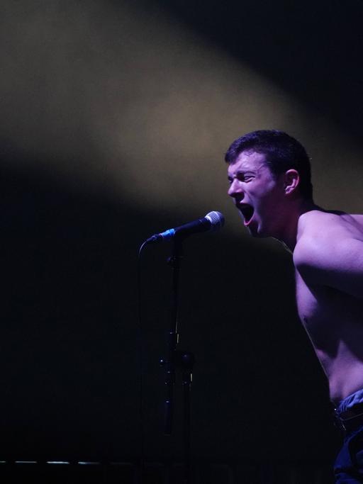 Charlie Steen von der Londoner Band Shame steht bei einem Live-Auftritt im September 2017 mit einem Band-Mitglied oben ohne auf der Bühne und singt kraftvoll in ein Mikrofon, die Arme nach hinten gestreckt.