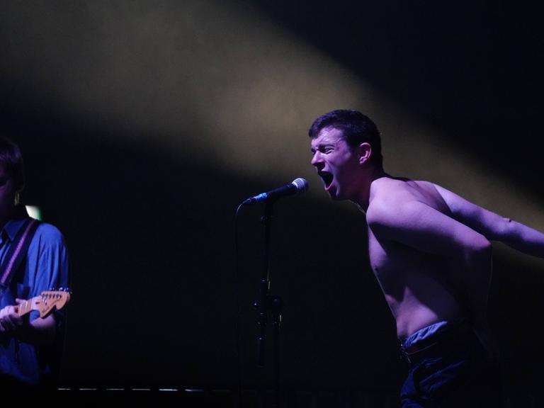 Charlie Steen von der Londoner Band Shame steht bei einem Live-Auftritt im September 2017 mit einem Band-Mitglied oben ohne auf der Bühne und singt kraftvoll in ein Mikrofon, die Arme nach hinten gestreckt.