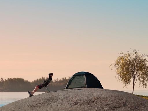 Ein Mann sitzt auf einem Steinhügel am Finnischen Meerbusen und blickt in die Ferne, hinter ihm steht ein kleines Zelt.