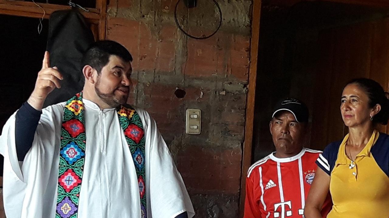 Der kolumbianische Priester Henry Ramirez trägt einen weißen Talar mit einer bunt bestickten Schärpe, er hält die rechte Hand in einer Zeigegeste erhoben, bei ihm stehen ein Mann im roten Hemd mit Schirmmütze und eine Frau in einem gelben Hemd mit ernsten Gesichtern.