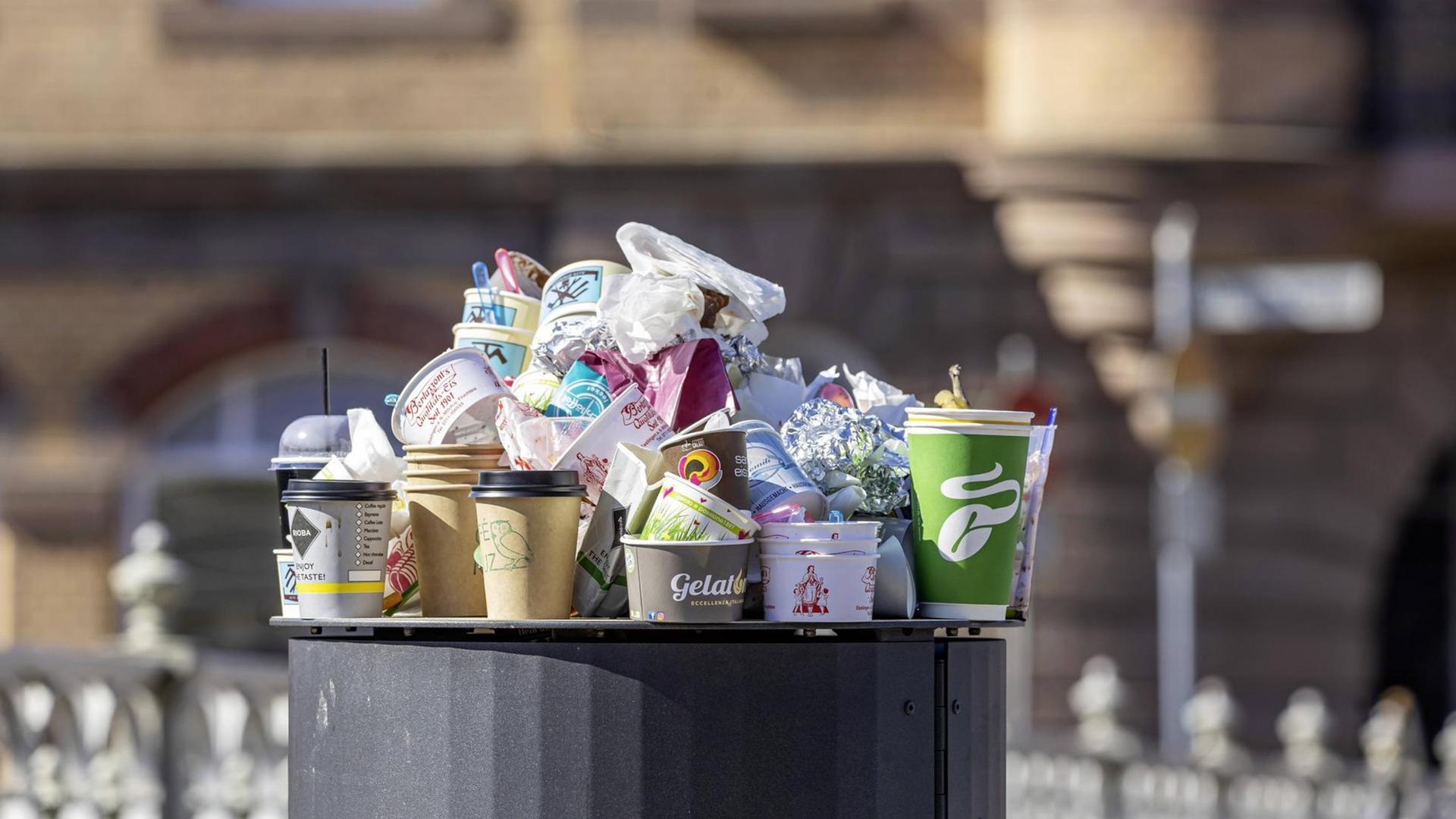 Leere Essens- und Getränke-Verpackungen stehen auf einem vollen Mülleimer. 