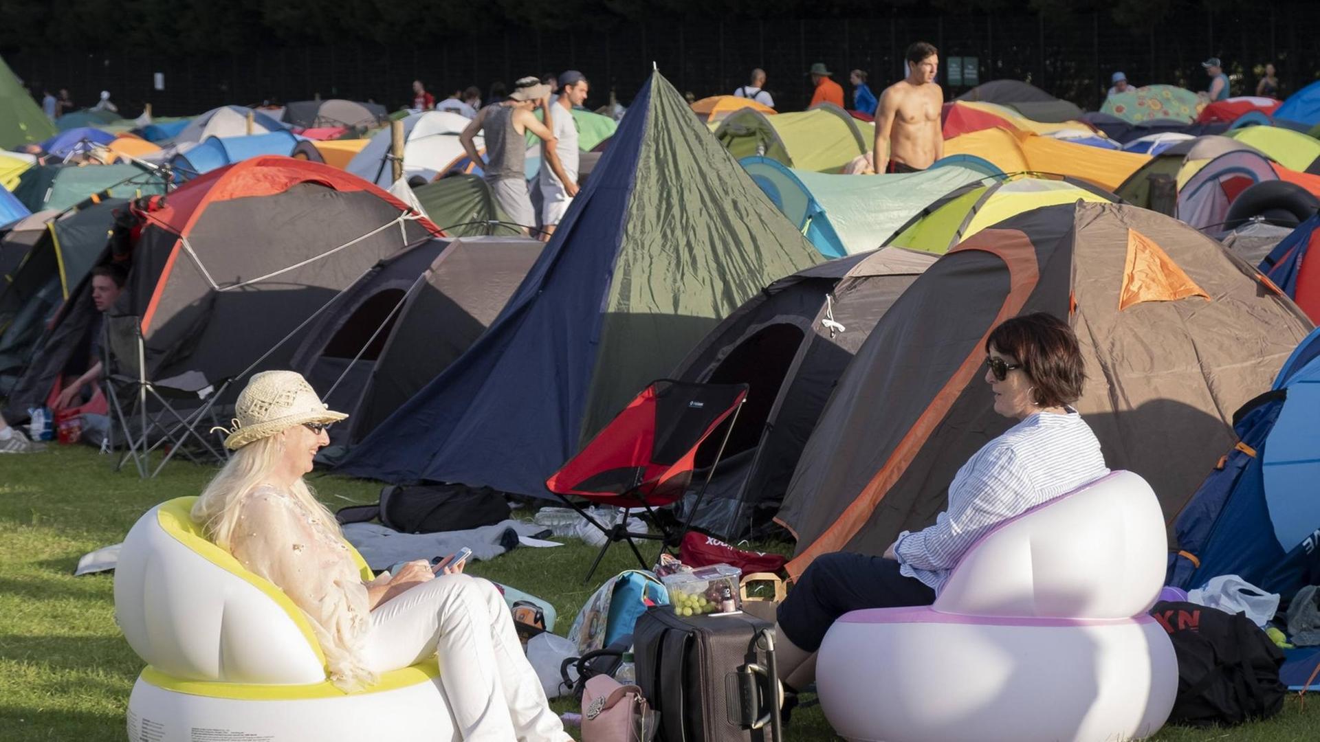 Zwei weibliche Tennis-Fans sitzen bequem in ihren mitgebrachten aufblasbaren Sitzmöbeln. Im Hintergrund: Zelte auf dem Camping-Platz von Wimbledon.