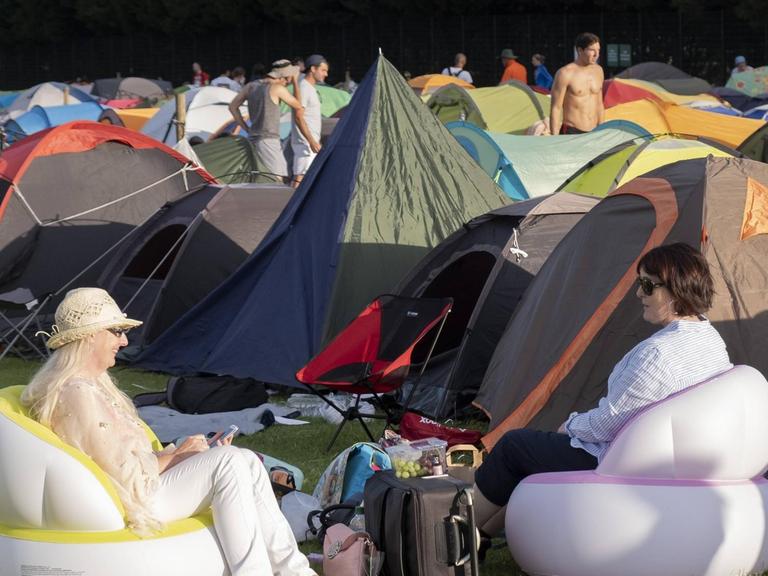 Zwei weibliche Tennis-Fans sitzen bequem in ihren mitgebrachten aufblasbaren Sitzmöbeln. Im Hintergrund: Zelte auf dem Camping-Platz von Wimbledon.