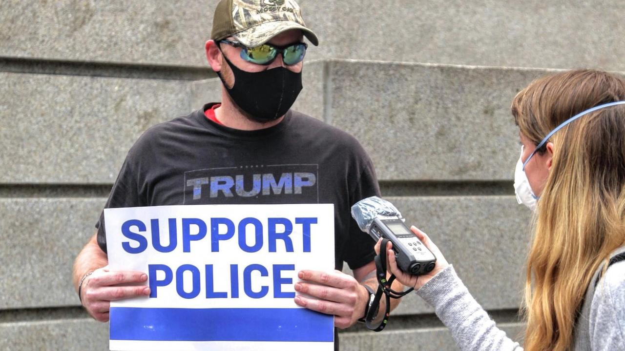 Ein Mann mit schwarzer Gesichtsmaske und in einem Trump T-Shirt wird interviewt. In seinen Händen hält er ein Transparent mit der Aufschrift "Support Police". Ein Mann mit schwarzer Gesichtsmaske und in einem Trump T-Shirt wird interviewt. In seinen Händen hält er ein Transparent mit der Aufschrift "Support Police".