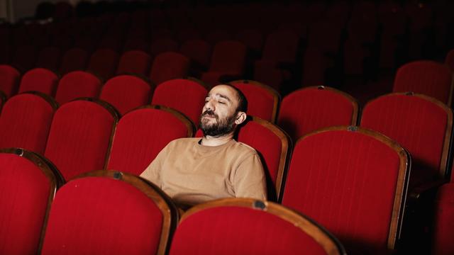 Ein Mann sitzt ganz alleine in einem Kinosaal mit roten Sesseln.