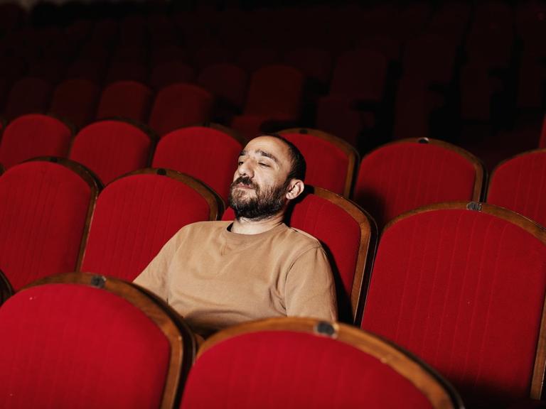 Ein Mann sitzt ganz alleine in einem Kinosaal mit roten Sesseln.