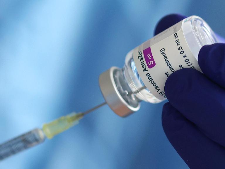 Ein blau behandschuhte Hand hält ein Fläschchen mit dem Covid-Impfstoff von AstraZeneca und präpariert eine Spritze.