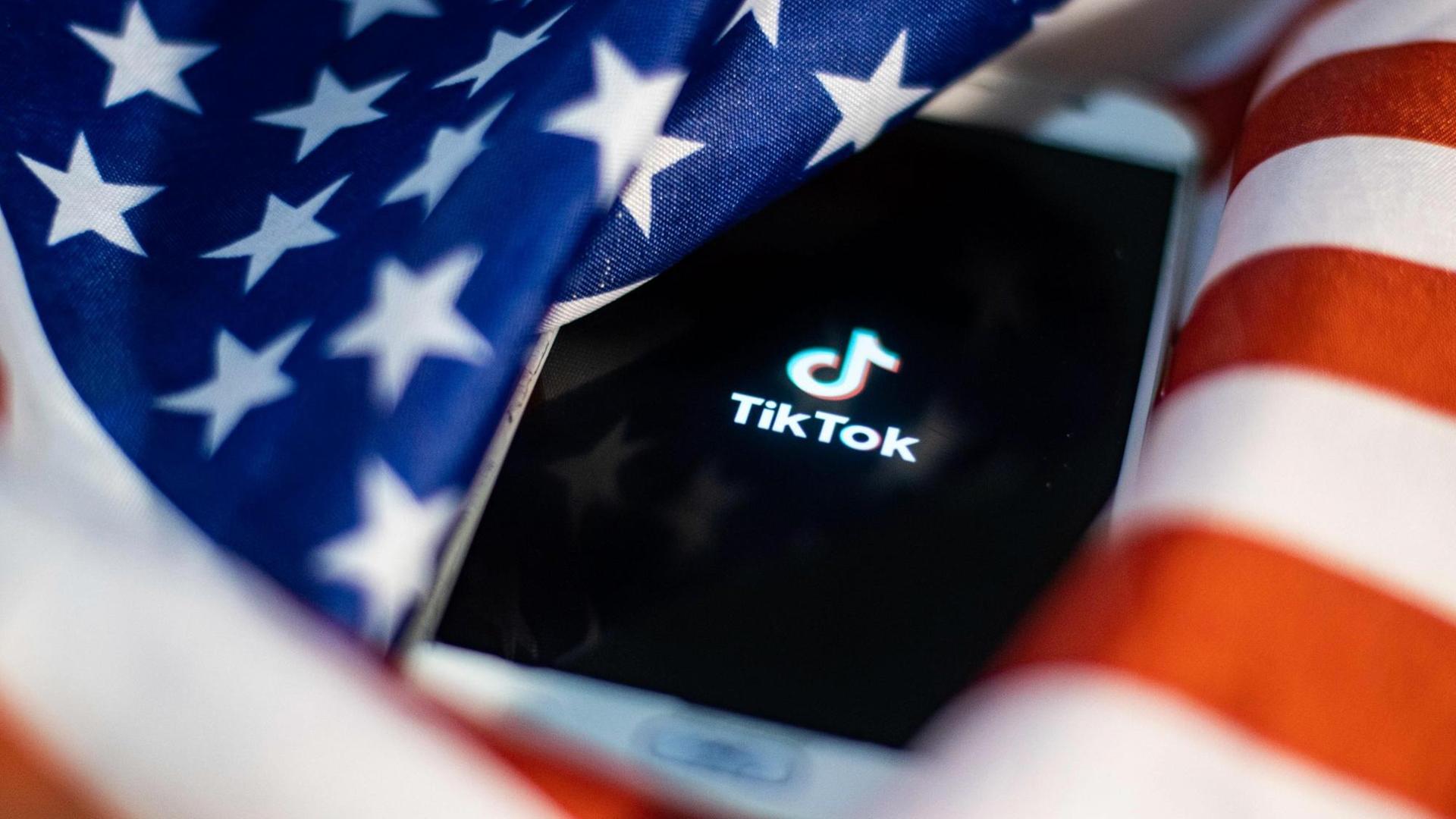 Ein Handy mit TikTok-Logo liegt auf einer US-Flagge. Ein Handy mit TikTok-Logo liegt auf einer US-Flagge.