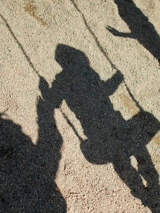 Der Schatten von einem Mann, einem schaukelnden Jungen und einem Mädchen fallen auf Sand auf einem Spielplatz.