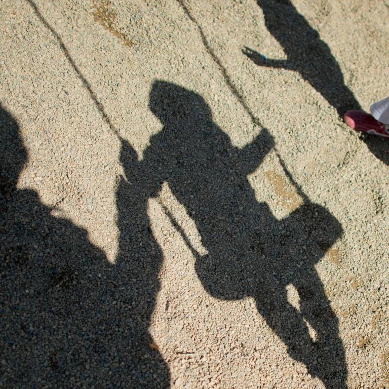 Der Schatten von einem Mann, einem schaukelnden Jungen und einem Mädchen fallen  auf Sand auf einem Spielplatz.