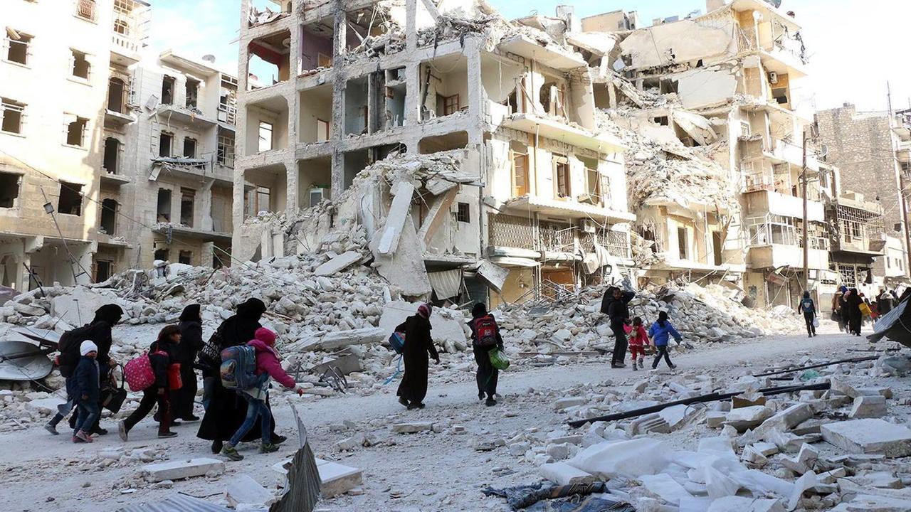 Sie sehen Menschen in Aleppo, die mit Gepäckstücken durch die zerstörten Straßen laufen. Im Hintergrund zerbombte Gebäude. Sie sehen Menschen in Aleppo, die mit Gepäckstücken durch die zerstörten Straßen laufen. Im Hintergrund zerbombte Gebäude.