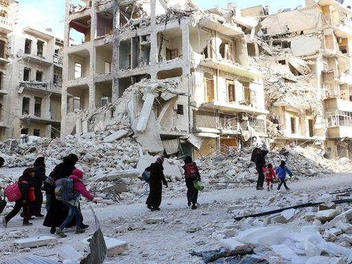 Sie sehen Menschen in Aleppo, die mit Gepäckstücken durch die zerstörten Straßen laufen. Im Hintergrund zerbombte Gebäude.