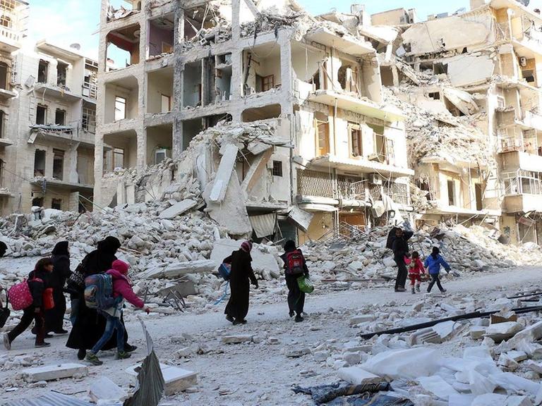 Sie sehen Menschen in Aleppo, die mit Gepäckstücken durch die zerstörten Straßen laufen. Im Hintergrund zerbombte Gebäude.