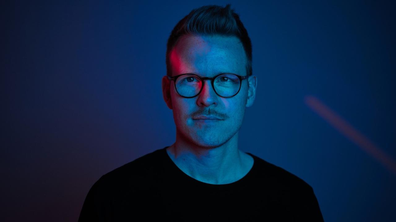 Ein junger Mann mit Brille, Oberlippenbart und Kurzhaarfrisur ist in blaues Licht getaucht im Porträt zu sehen. Er blickt den Betrachter direkt an. Ein junger Mann mit Brille, Oberlippenbart und Kurzhaarfrisur ist in blaues Licht getaucht im Porträt zu sehen. Er blickt den Betrachter direkt an.