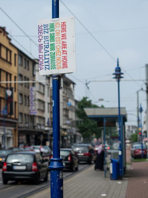 Ein Schild "Hier sind wir zuhause" in verschiedenen Sprachen hängt am 12.08.2015 in Duisburg (Nordrhein-Westfalen) auf der Straße. Händlern brechen die Geschäfte weg, Familienclans reklamieren die Gegend für sich.
