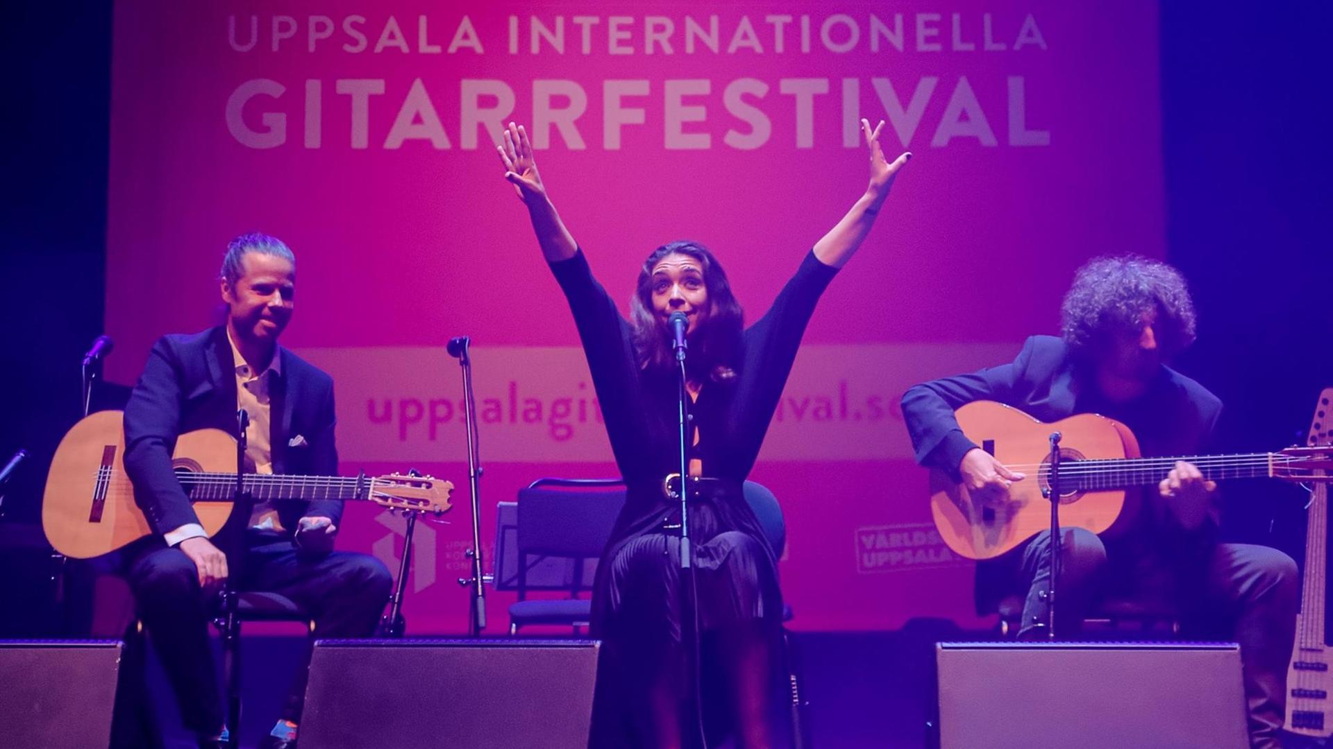 Drei Künstler sitzen auf der Bühne, die Sängerin in der Mitte, die beiden Gitarristen ihr zu Seite. Sie streckt beide Arme in die Höhe, im Hintergrund das Festivalplakat auf pinkfarbenem Grund: Uppsala Internationella Gitarrfestival Drei Künstler sitzen auf der Bühne, die Sängerin in der Mitte, die beiden Gitarristen ihr zu Seite. Sie streckt beide Arme in die Höhe, im Hintergrund das Festivalplakat auf pinkfarbenem Grund: Uppsala Internationella Gitarrfestival