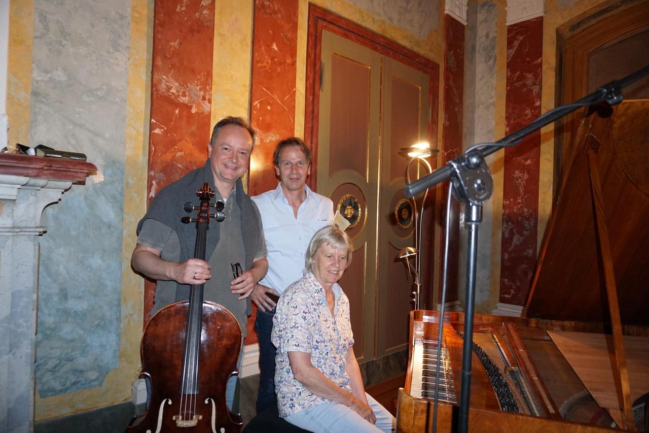 Der Cellist mit Cello in der Hand, der Moderator mit Noten unter dem Arm und die Pianistin neben dem Hammerklavier auf einem Hocker schauen in die Kamera. Der Cellist mit Cello in der Hand, der Moderator mit Noten unter dem Arm und die Pianistin neben dem Hammerklavier auf einem Hocker schauen in die Kamera.