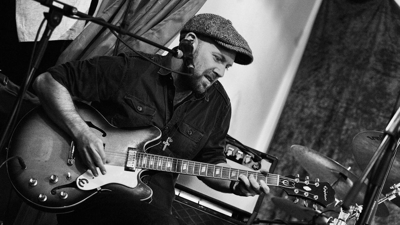 Mann mit Schirmmütze sitzt mit Gitarre auf einem Hocker und spielt. Mann mit Schirmmütze sitzt mit Gitarre auf einem Hocker und spielt.